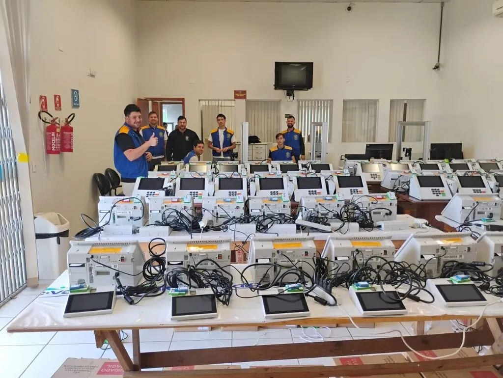 Urnas eletrônicas dispostas em mesas no Fórum Eleitoral de Pinhão, com cabos e materiais de preparação. Integrantes da equipe da Justiça Eleitoral aparecem ao fundo da sala.