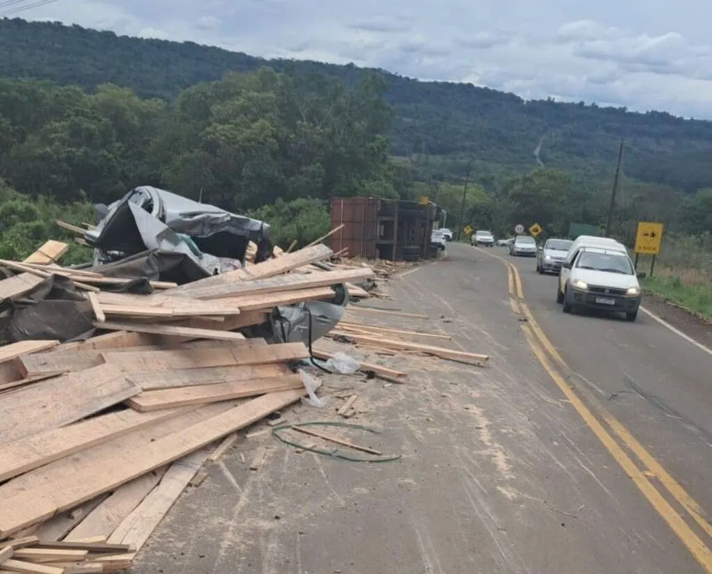 Tábuas de madeira espalhadas sobre uma das faixas da rodovia, junto a um automóvel com a carroceria destruída. Ao fundo, um veículo de carga está tombado, enquanto carros passam pela faixa