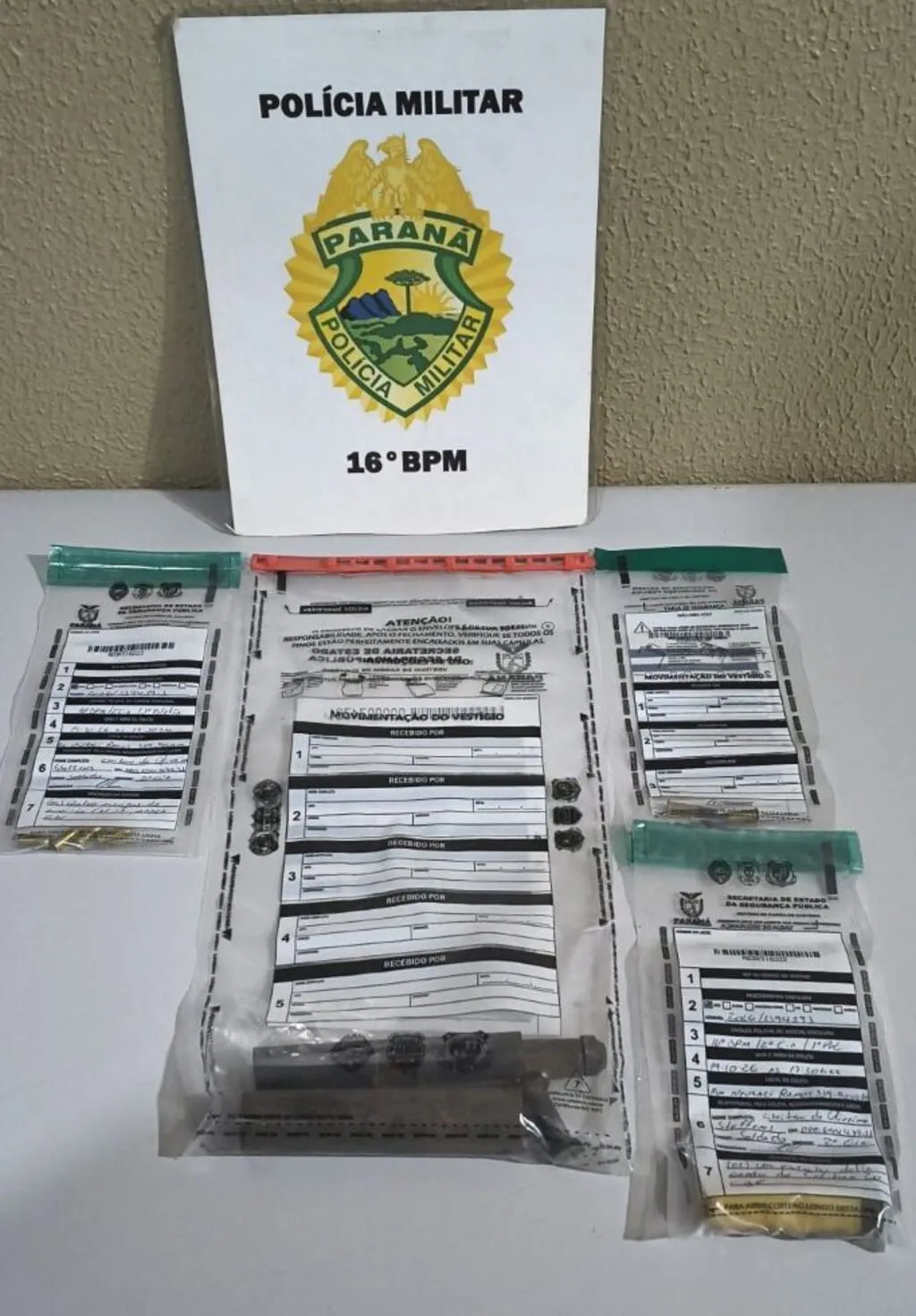 Arma artesanal, munições e estojos acondicionados em embalagens de evidências sobre uma mesa, diante de uma placa com o brasão da Polícia Militar do Paraná e a identificação do 16º BPM.