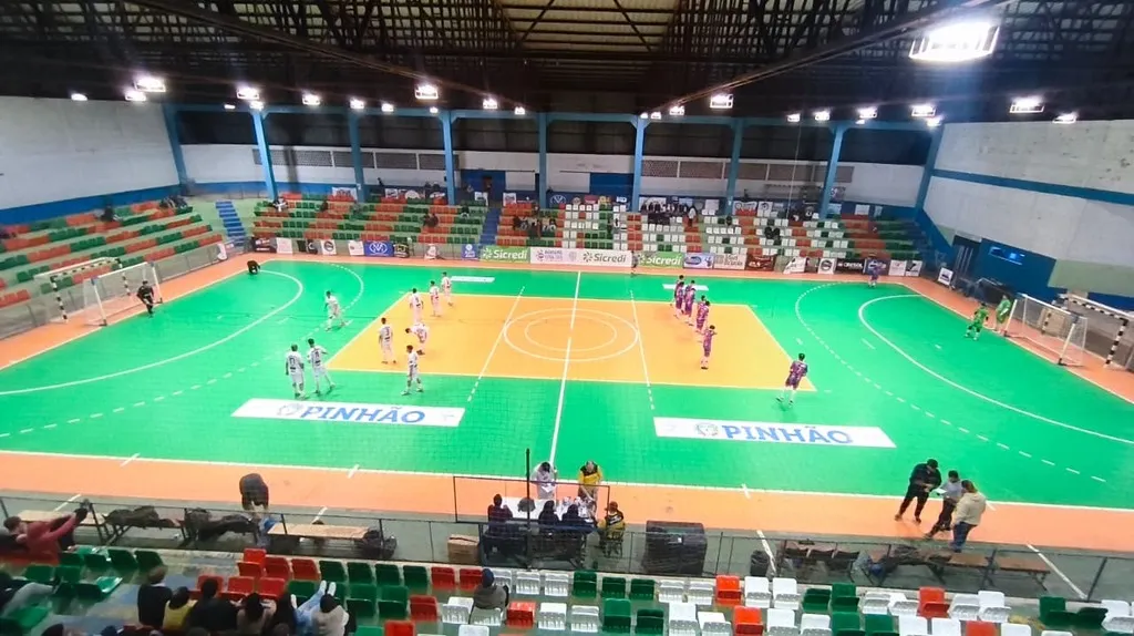 Vista ampla da quadra do Ginásio XV de Dezembro, em Pinhão, com jogadores de uniformes brancos e roxos, equipe de arbitragem e espectadores nas arquibancadas durante a terceira rodada do Campeonato Municipal de Futsal.