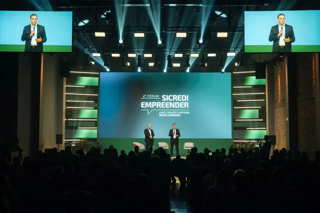 Dois homens estão no palco diante do público no 2º Fórum Sicredi Empreender. Um deles fala ao microfone, enquanto o telão central exibe o nome do evento e telas laterais mostram sua imagem ampliada.